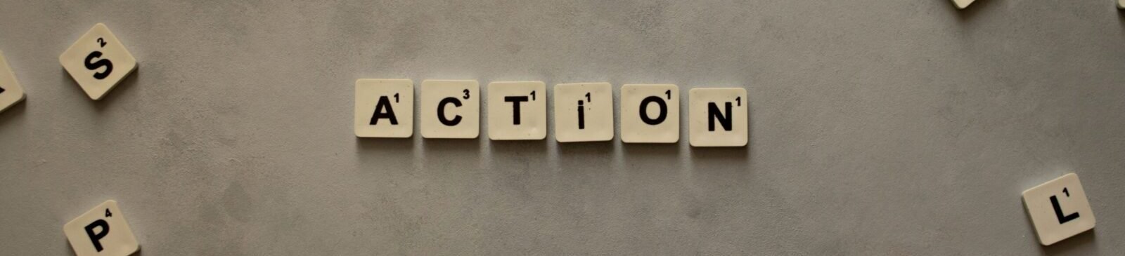 Flat lay of Scrabble tiles spelling 'Action' on a gray background, surrounded by random letters.